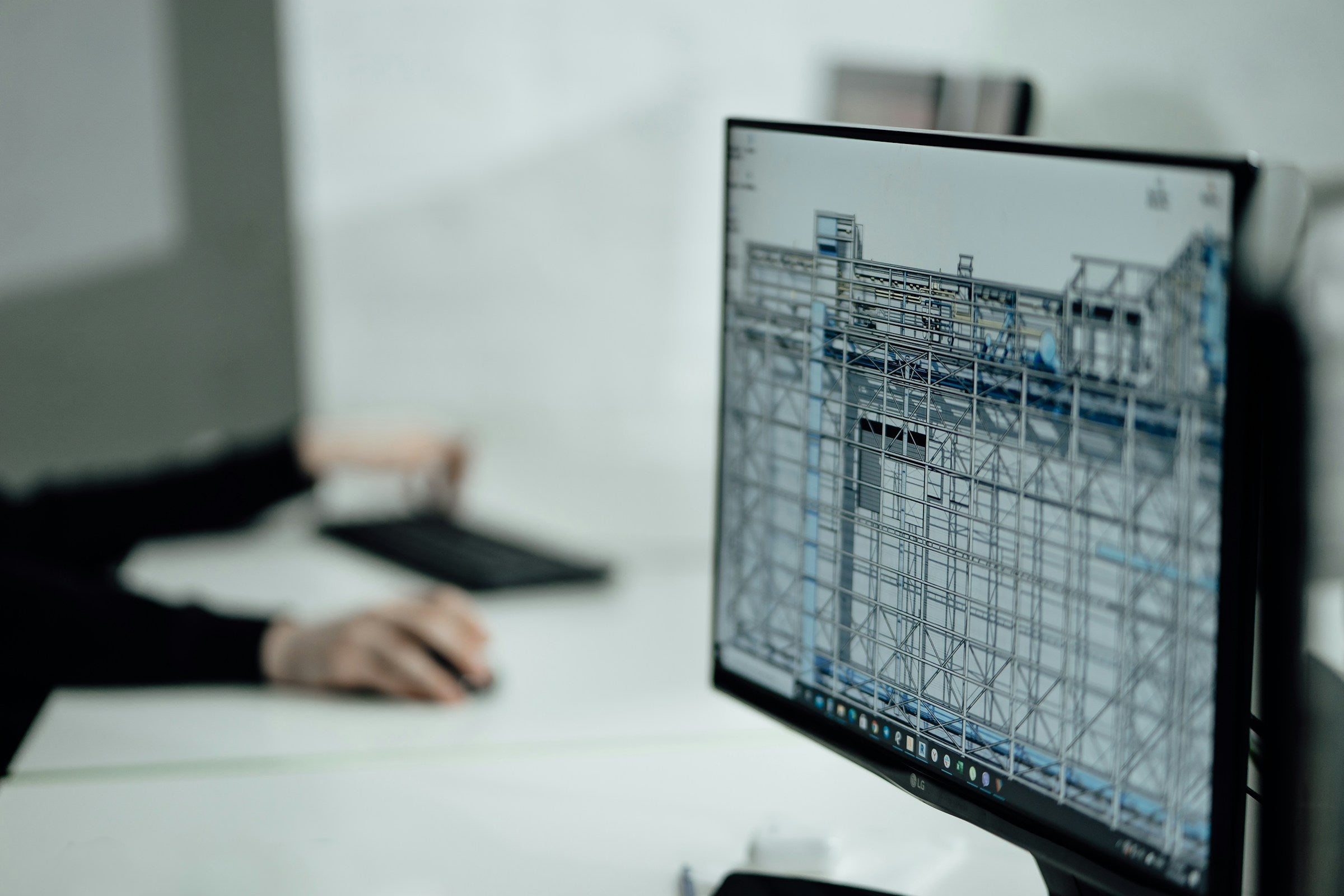 A computer monitor on a white desk displaying a blue and white architectural or structural CAD model, with a person's hands and arms blurred in the background using a keyboard and mouse.
